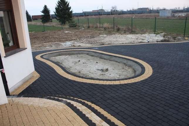 Układanie kostki brukowej w kolorze grafitowym z obrzeżem w kolorze piaskowym, przygotowany teren pod oczko wodne z obrzeżem z kostki, widok na ogród z drzewami i linią kolejową w oddali.