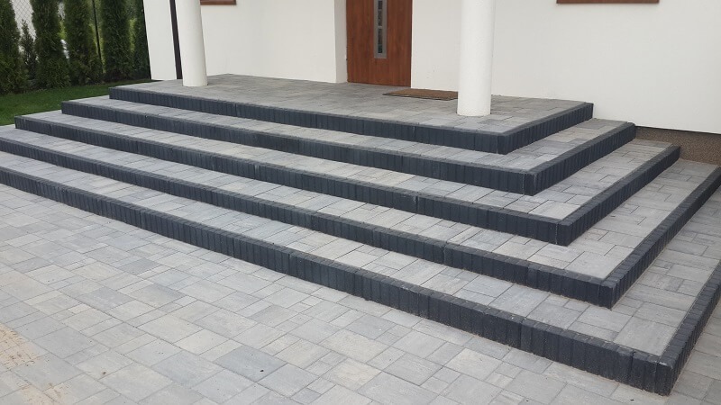 Schody wejściowe do domu wyłożone szarą kostką brukową z ciemnoszarym obrzeżem, widok z dołu, trawnik w tle.