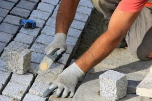 Pracownik w rękawicach roboczych układa kostkę granitową, wyrównując ją gumowym młotkiem. Widoczny fragment chodnika z szarej kostki oraz narzędzia brukarskie.