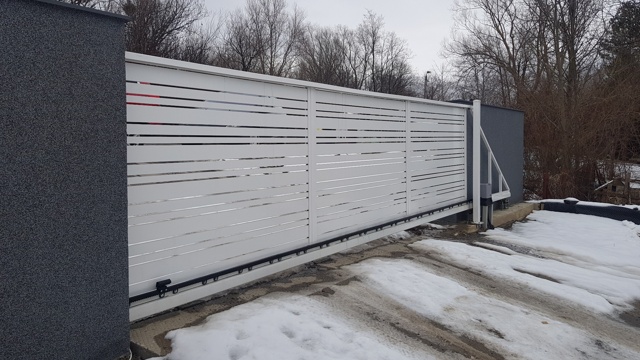 Nowoczesna, przesuwna brama w kolorze białym z poziomymi szczelinami, zamontowana na posesji z resztkami śniegu, widoczny mechanizm automatyczny i fragment ściany w kolorze grafitowym.