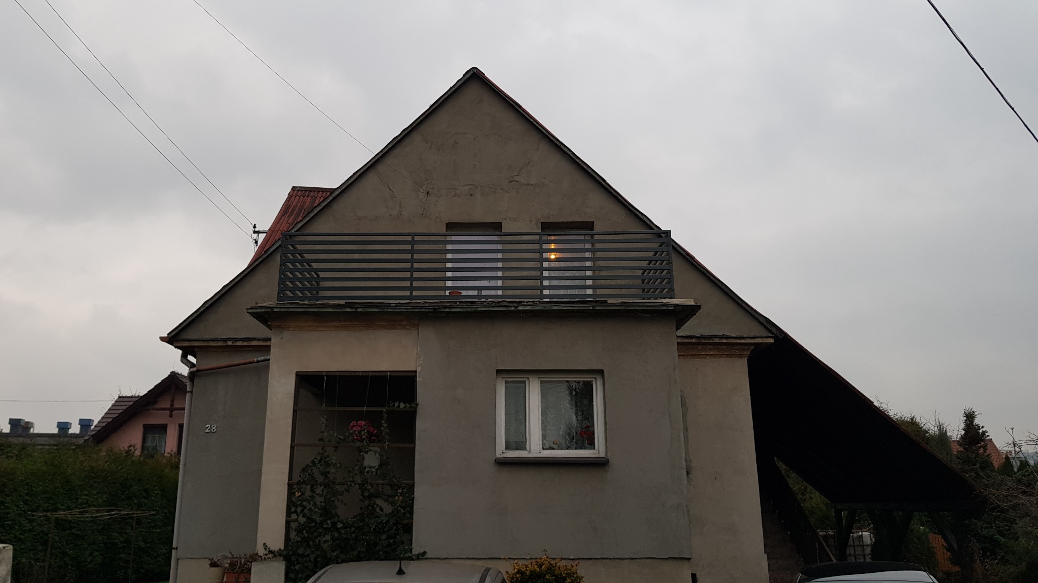 Szary dom z balkonem z metalową balustradą w poziome paski, widok z dołu na pochmurne niebo, w oknach odbija się światło.