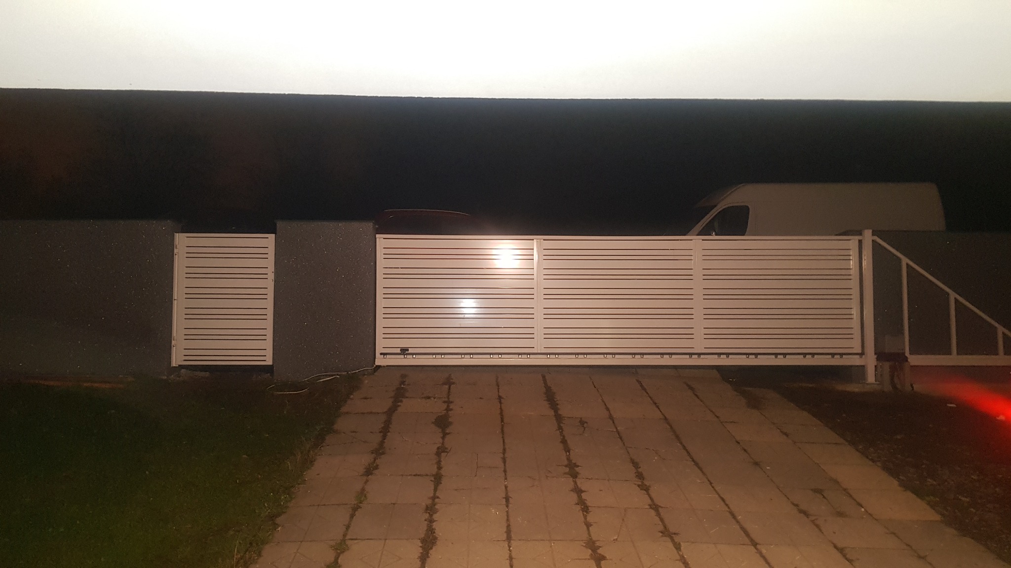 Nowoczesna, przesuwna brama wjazdowa z poziomymi panelami, oświetlona od góry, z widocznym fragmentem ogrodzenia i podjazdu z kostki brukowej w tle zaparkowany biały samochód dostawczy.