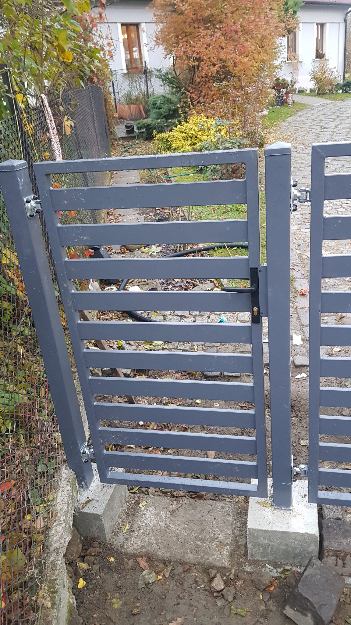 Szara, pozioma brama lamelowa z czarną klamką i zamkiem, zamontowana na betonowych słupkach w ogrodzeniu posesji z widocznym domem i jesienną roślinnością w tle.