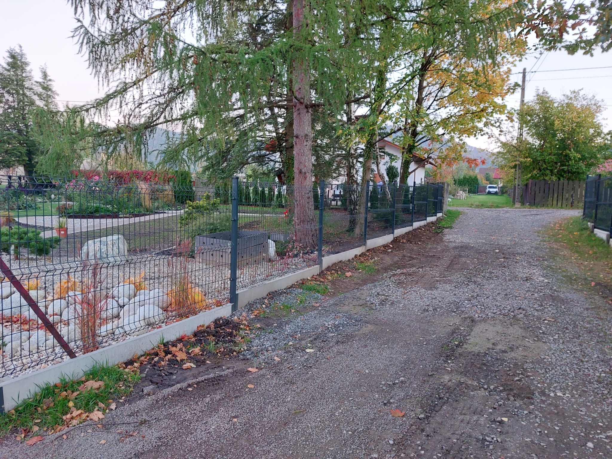 Zielony panelowy system ogrodzeniowy z betonową podmurówką, widok z drogi szutrowej, z drzewami i ogrodem w tle.