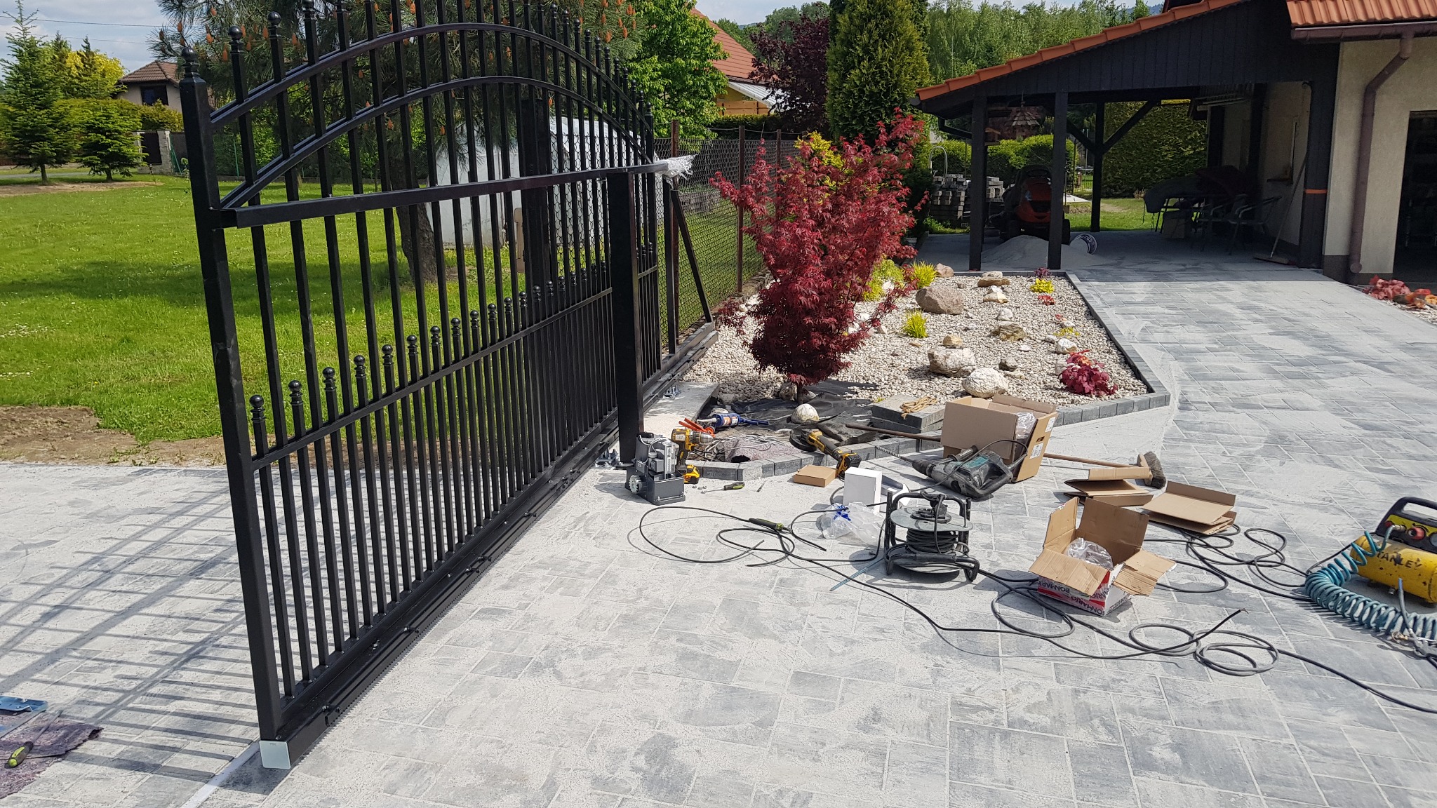 Czarna, metalowa brama przesuwna z ozdobnymi elementami, w trakcie montażu na posesji z kostki brukowej, widoczne narzędzia i materiały montażowe.