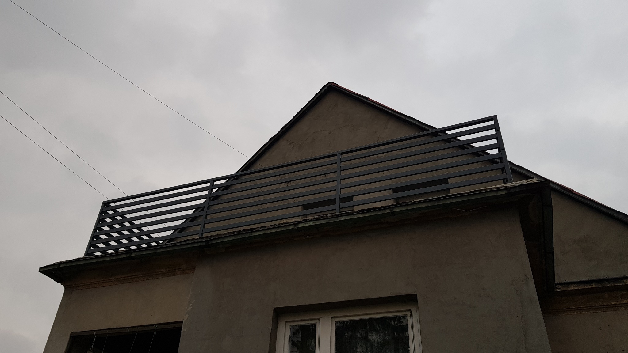 Szare, poziome ogrodzenie z metalowych profili na dachu budynku z szarą elewacją, widoczne przewody elektryczne na tle pochmurnego nieba.