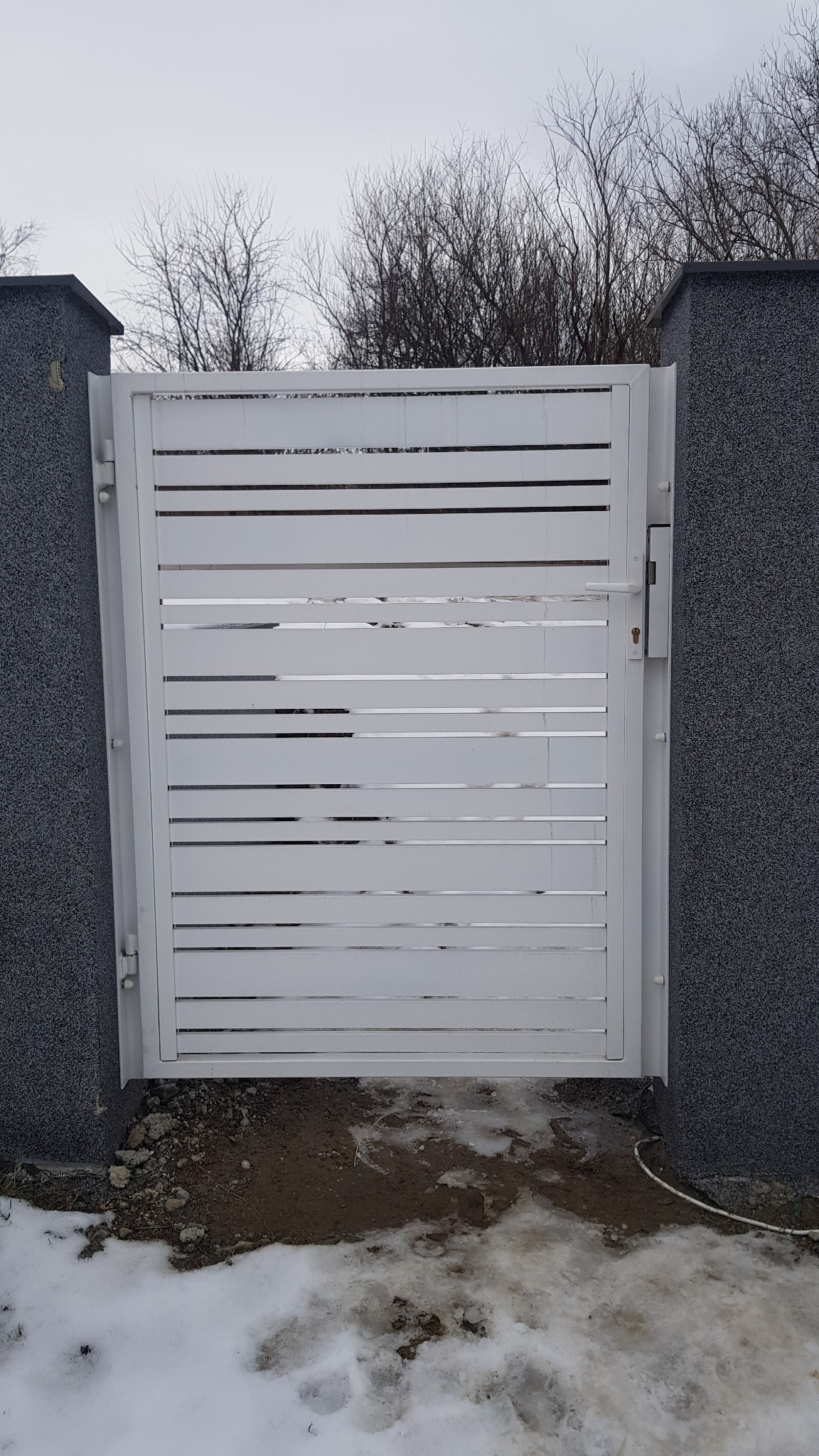 Nowoczesna, biała brama z poziomymi szczeblami, zamontowana między dwoma szarymi, teksturowanymi słupkami. Ziemia pokryta resztkami śniegu, w tle nagie drzewa.