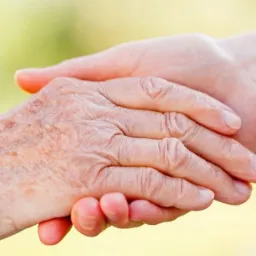 Dłoń osoby starszej trzymana przez dłoń młodszą, ukazująca troskę i wsparcie na rozmytym, jasnym tle.