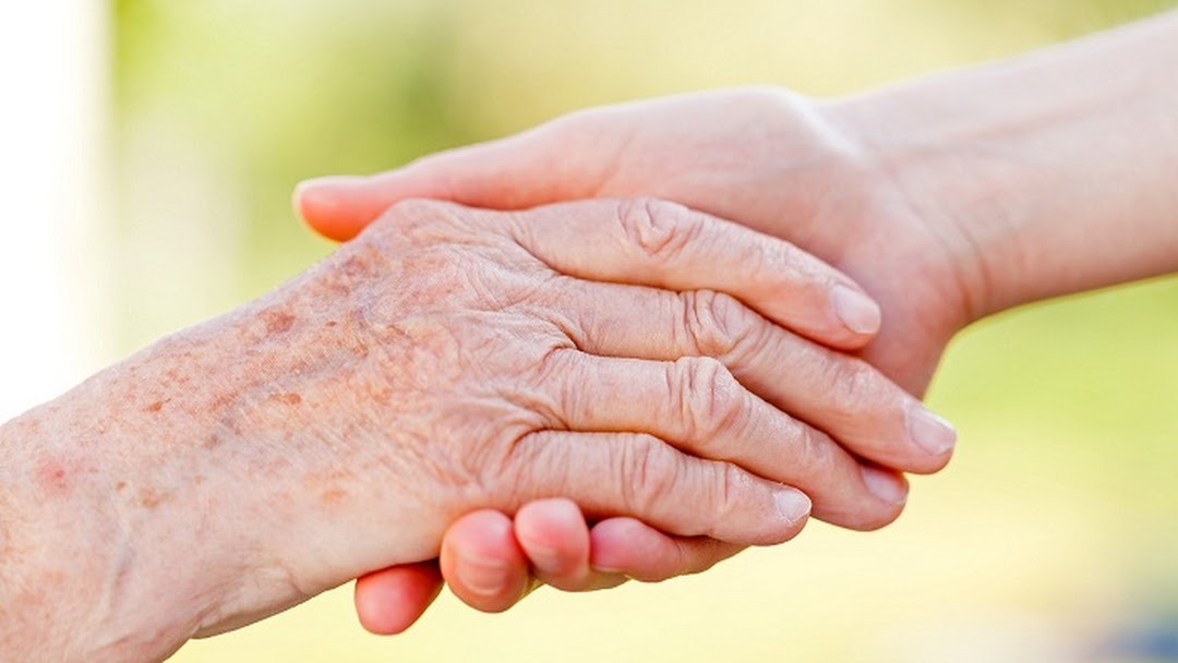 Dłoń osoby starszej trzymana przez dłoń młodszą, ukazująca troskę i wsparcie na rozmytym, jasnym tle.