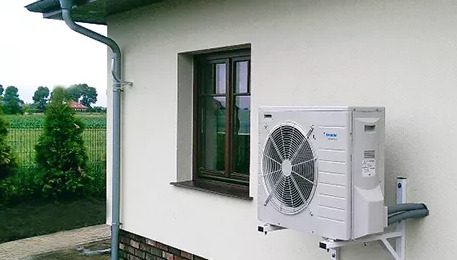 Zewnętrzna jednostka klimatyzacji zamontowana na białej ścianie domu jednorodzinnego, widoczny odpływ rynny i okno z brązową ramą.