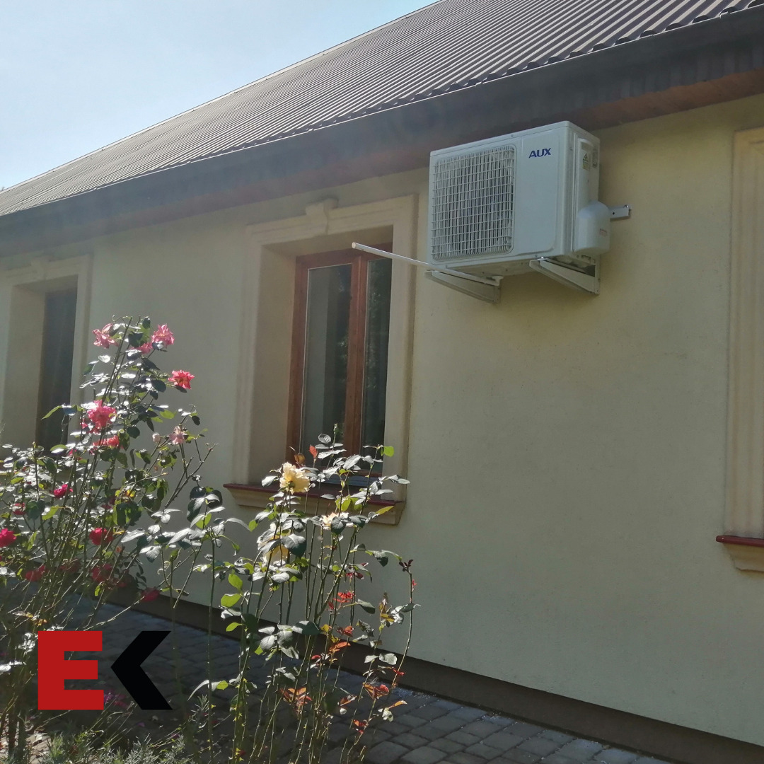 Biały klimatyzator AUX zamontowany na elewacji domu z beżowym tynkiem, widoczny spod dachu z blachy trapezowej, na tle kwitnących krzewów róż.