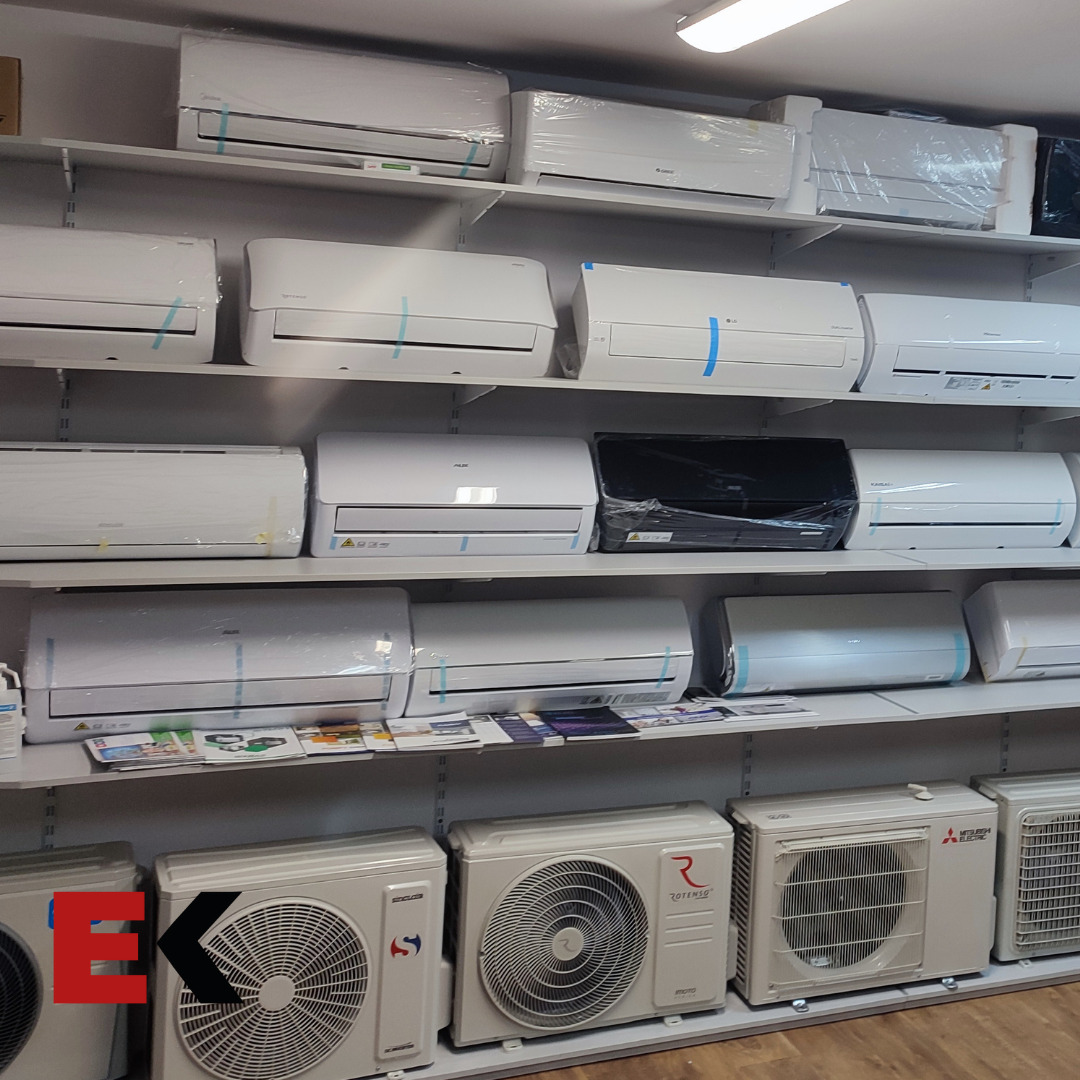 Ekspozycja klimatyzatorów różnych marek na półkach sklepowych, widoczne jednostki wewnętrzne i zewnętrzne, niektóre jeszcze w folii ochronnej.