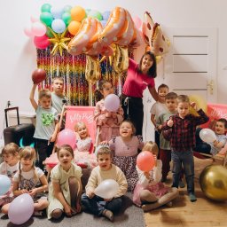 LollyPolly - Uśmiechnięta animatorka w otoczeniu dzieci z balonami na tle dekoracji urodzinowej. Radosna atmosfera imprezy dziecięcej z motywem przewodnim.