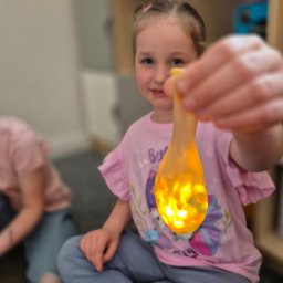 LollyPolly - Dziewczynka trzymająca świecący balon z brokatem, siedząc na podłodze. W tle regał z zabawkami. Kreatywna zabawa dla dzieci na imprezie urodzinowej.