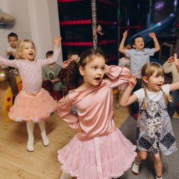 LollyPolly - Rozbawione dzieci w różowych strojach tańczą w sali zabaw z kolorową konstrukcją do wspinaczki w tle. Dziewczynka z brokatem na twarzy w centrum kadru.