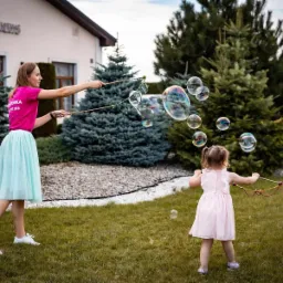 Kobieta w różowej koszulce i turkusowej spódnicy puszcza bańki mydlane, a mała dziewczynka w różowej sukience próbuje je łapać na trawniku przed budynkiem.