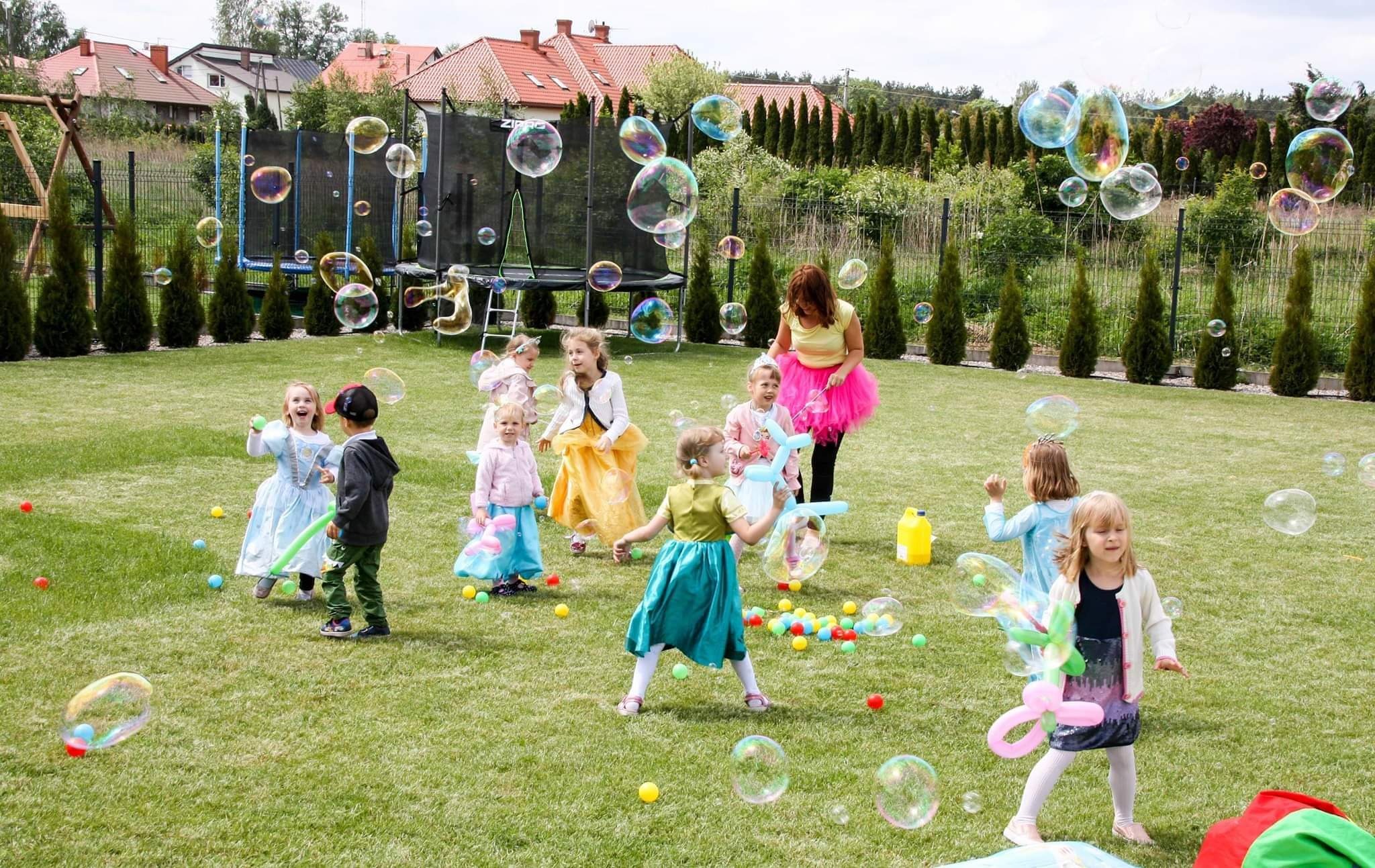 Animator w różowej tiulowej spódnicy dmucha bańki mydlane dla grupy dzieci w kostiumach na trawniku, w tle trampolina i domy.