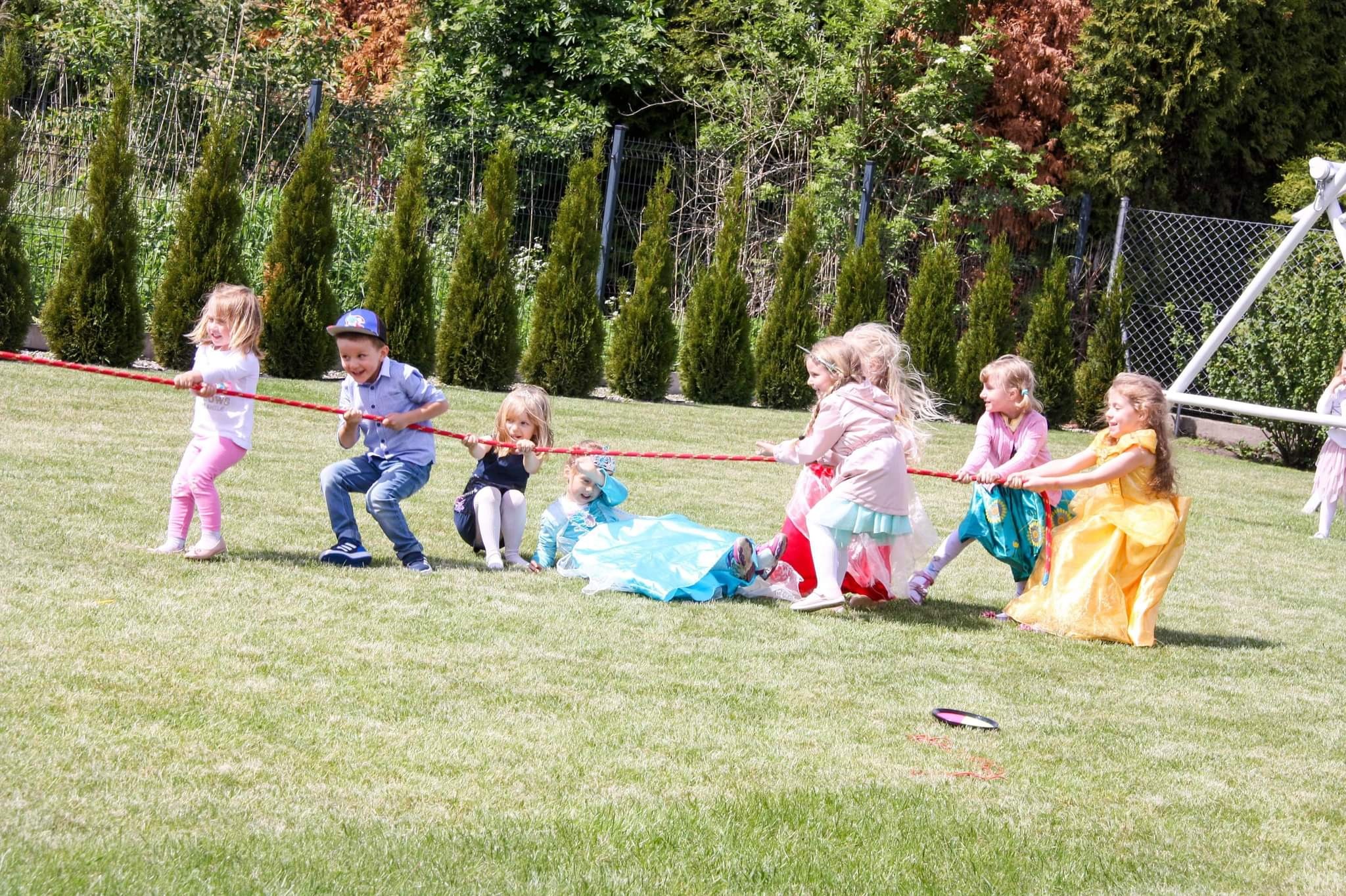 Grupa dzieci, niektóre w kostiumach księżniczek, bierze udział w przeciąganiu liny na trawniku podczas imprezy plenerowej, z tujami i ogrodzeniem w tle.