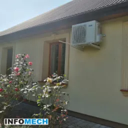 Biały klimatyzator AUX zamontowany na elewacji domu z kremową fasadą, widoczny z boku, z krzewem róż w pierwszym planie.