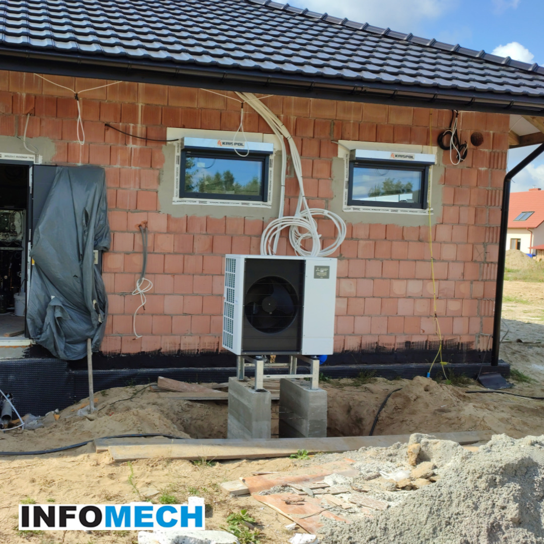 Biała pompa ciepła zamontowana na betonowych podstawach przy ceglanym budynku w trakcie budowy, widoczne przewody i okna z roletami zewnętrznymi.