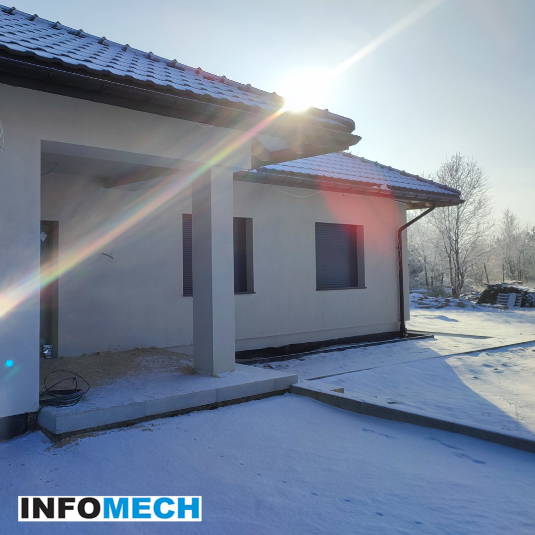 Nowy, energooszczędny dom jednorodzinny w zimowej scenerii, z widocznym fragmentem dachu pokrytego dachówką, oknami z roletami zewnętrznymi i elementami wykończeniowymi w śniegu, oświetlony...