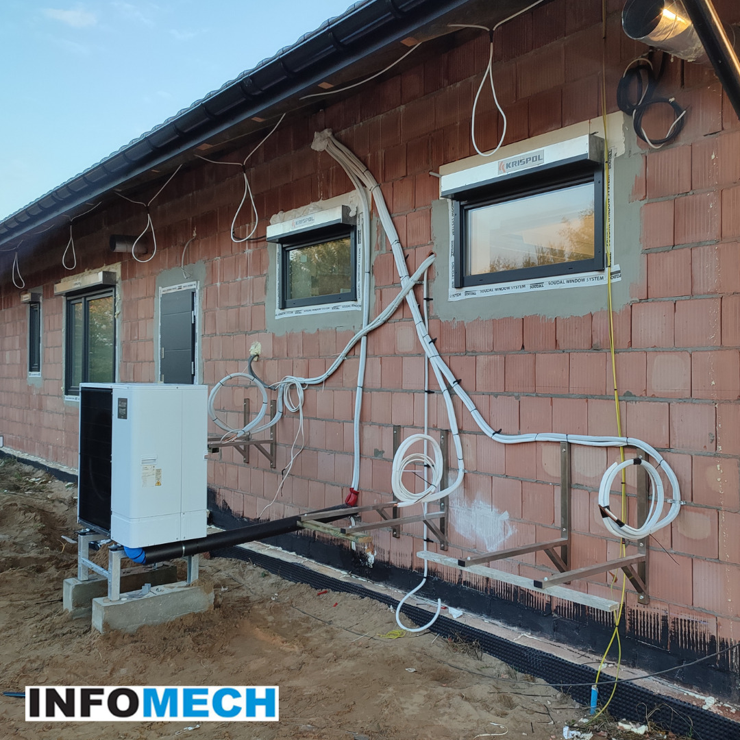 Zewnętrzna jednostka grzewcza zamontowana na metalowej konstrukcji przy ścianie budynku z czerwonej cegły. Widoczne przewody i rury prowadzone po elewacji, okna z roletami zewnętrznymi i logo firmy...