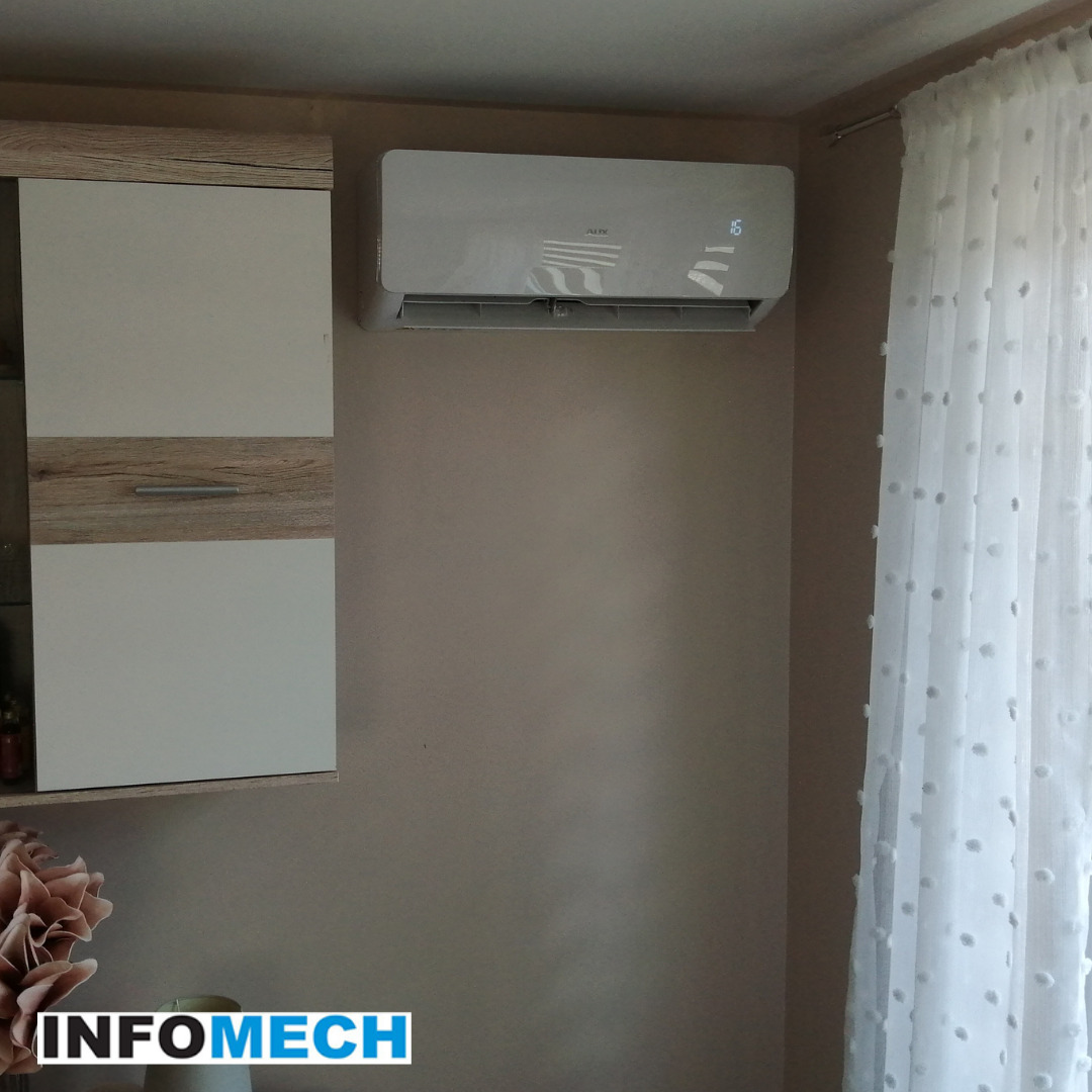 Biały klimatyzator ścienny marki AUX zamontowany w pokoju, obok szafki wiszącej i zasłony w kropki.