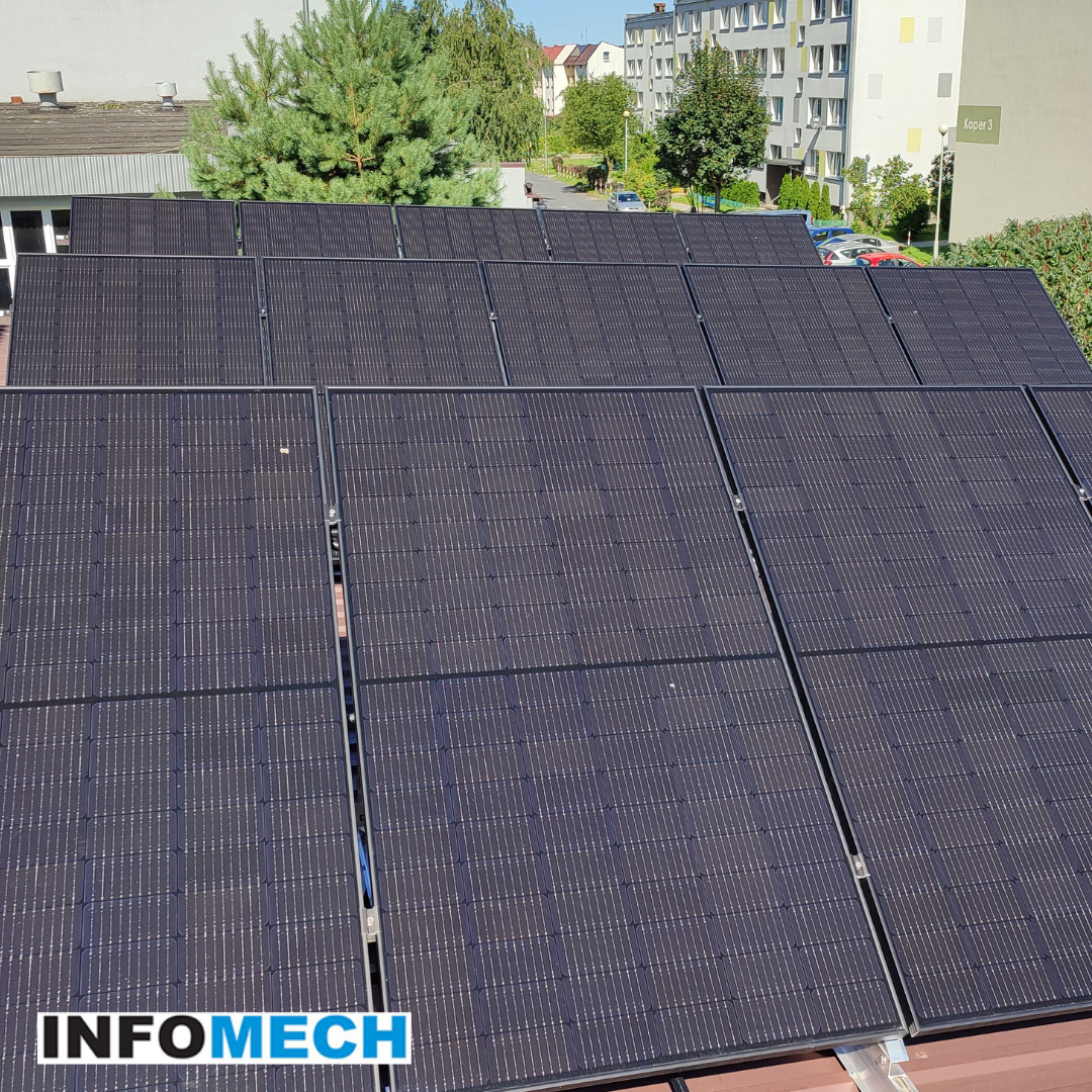 Instalacja paneli słonecznych na dachu budynku, widok z góry, z widocznymi drzewami i budynkami w tle. Na pierwszym planie widoczne panele z logo firmy w lewym dolnym rogu.