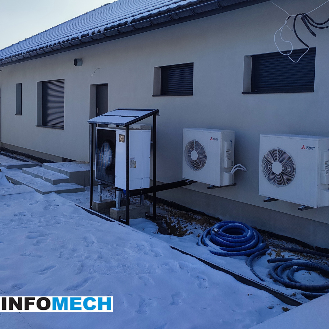 Zewnętrzna instalacja pompy ciepła Mitsubishi Electric z dwoma jednostkami zewnętrznymi zamontowanymi na ścianie budynku, widoczne niebieskie węże i śnieg na ziemi.