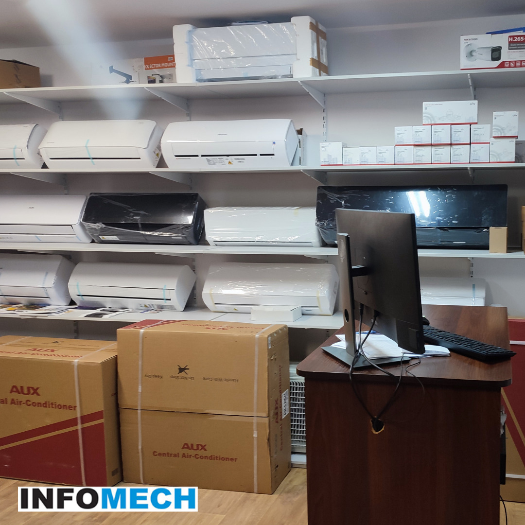 Salon z klimatyzatorami: Rzędy białych i czarnych jednostek wewnętrznych klimatyzacji na półkach, z kartonami klimatyzatorów AUX na podłodze i komputerem na biurku na pierwszym planie.
