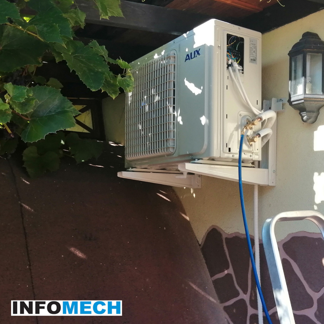 Zamontowana zewnętrzna jednostka klimatyzacji AUX na wspornikach, widoczne podłączenia rur miedzianych i przewód, obok lampa zewnętrzna i fragment zieleni.