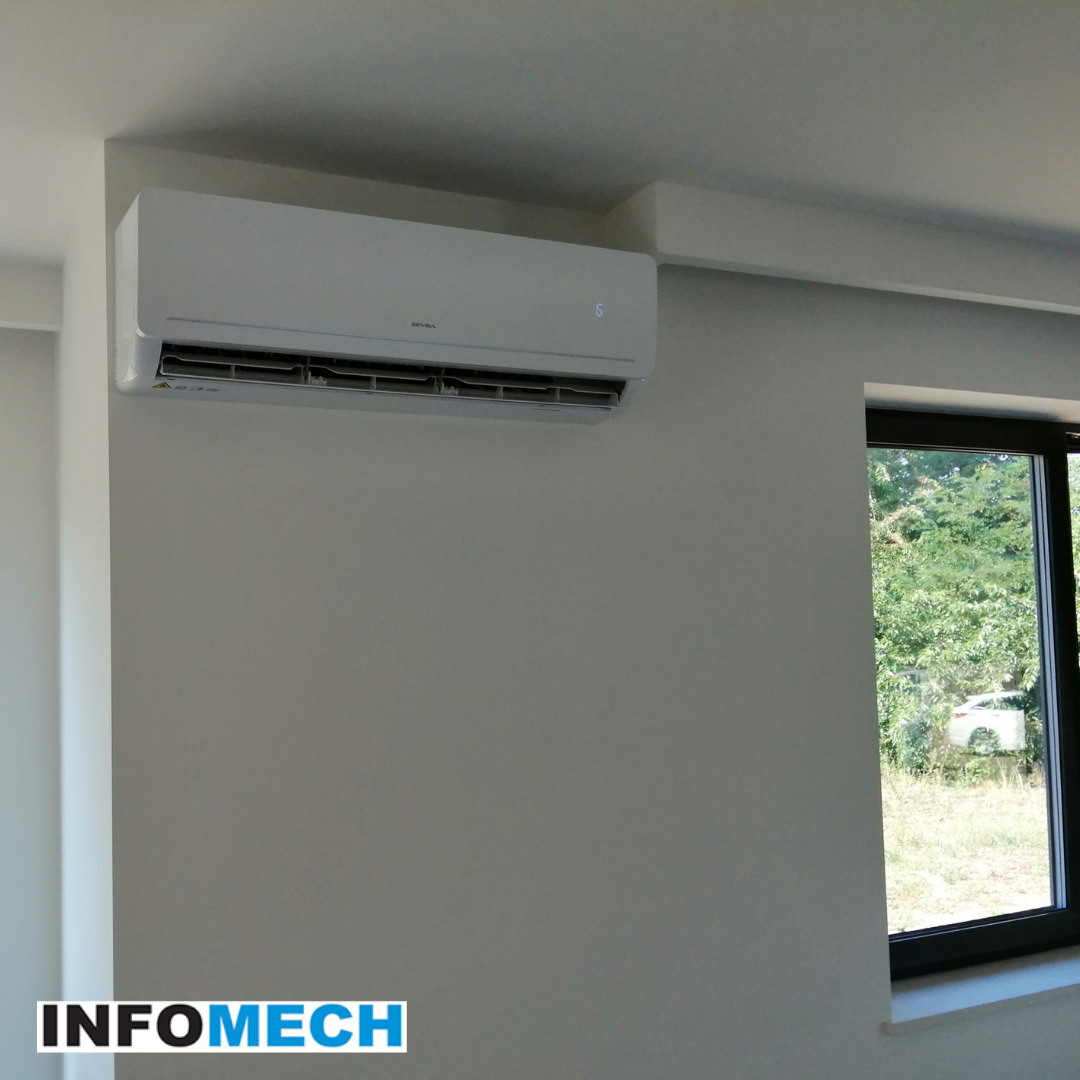 Biała jednostka klimatyzacyjna na ścianie w jasnym pokoju, widok z okna na zewnątrz, logo firmy w dolnej części zdjęcia.