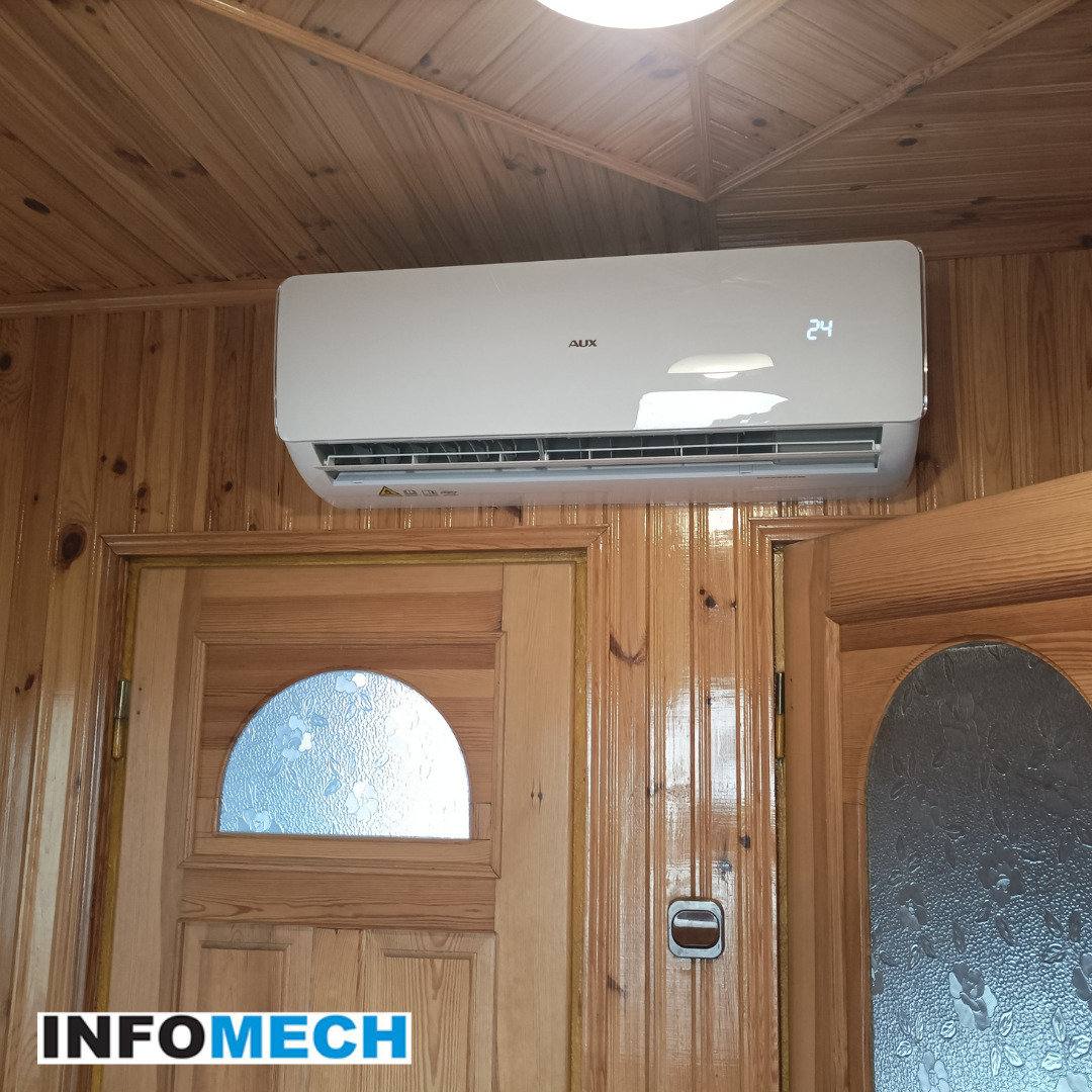 Biały klimatyzator AUX zamontowany nad drewnianymi drzwiami z półokrągłym przeszkleniem w pomieszczeniu wykończonym boazerią, widoczny wyświetlacz temperatury 24 stopnie.