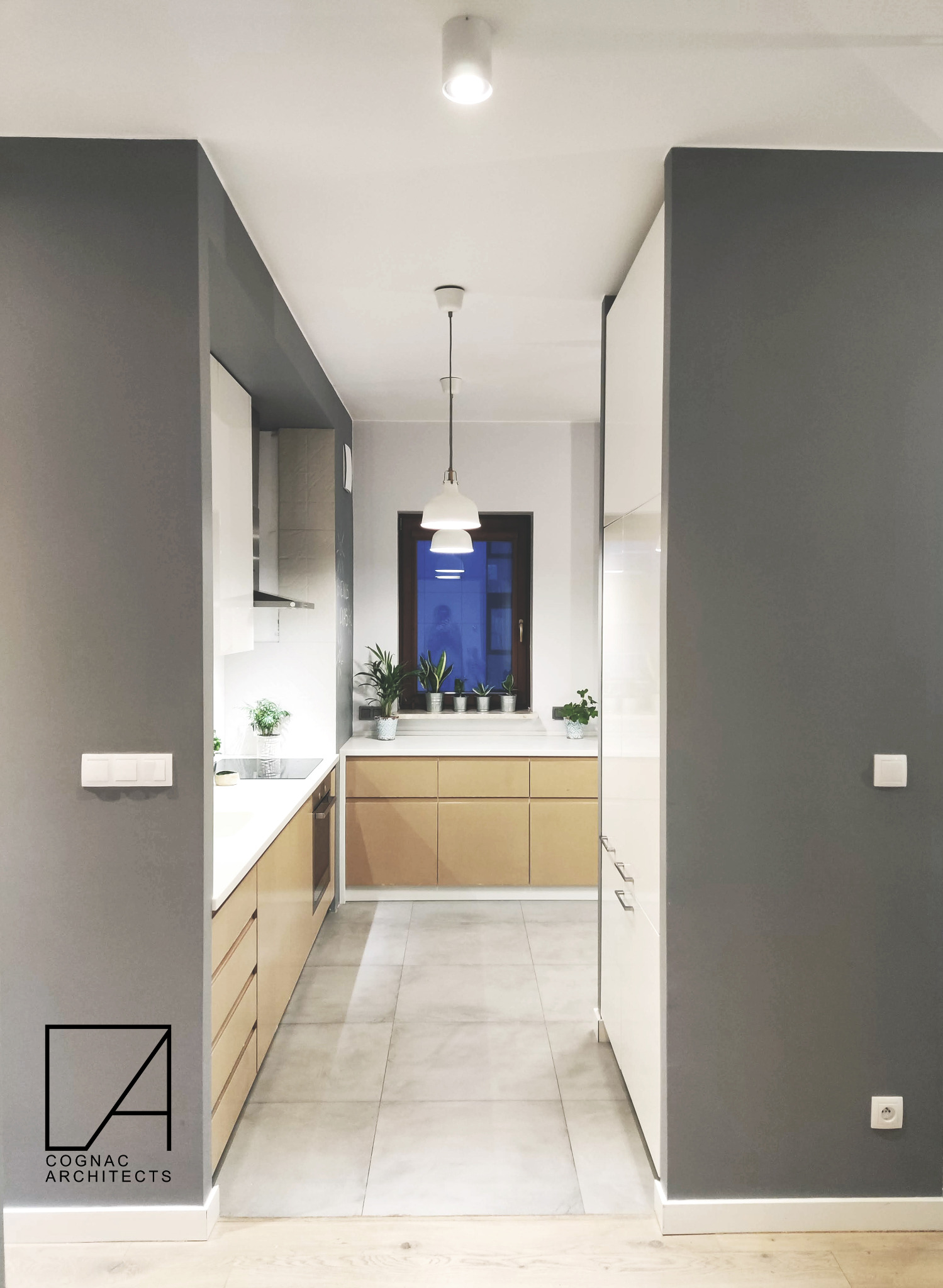 Wnętrze nowoczesnej kuchni w odcieniach szarości, bieli i naturalnego drewna, z trzema lampami wiszącymi nad blatem roboczym i rzędem doniczek z roślinami przy oknie. Logo Cognac Architects w lewym...