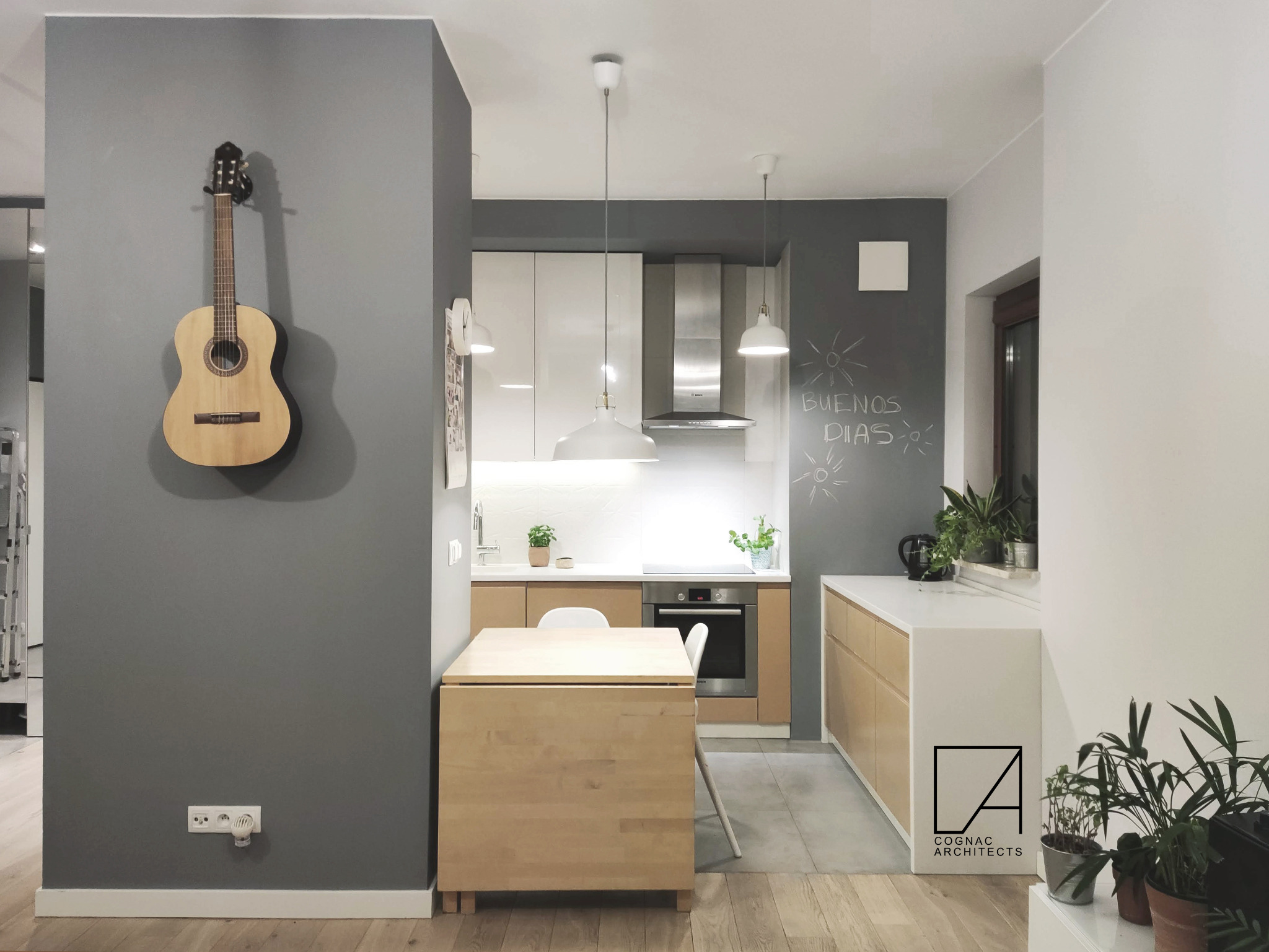 Wnętrze kuchni z gitarą akustyczną wiszącą na szarej ścianie, składany drewniany stół z krzesłem, białe i drewniane szafki kuchenne, okap ze stali nierdzewnej, tablica kredowa z napisem 'Buenos...