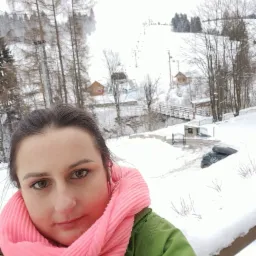 Kobieta w różowym szaliku i zielonej kurtce robiąca selfie na tle zimowego krajobrazu z zalesionymi wzgórzami i wyciągiem narciarskim w oddali.