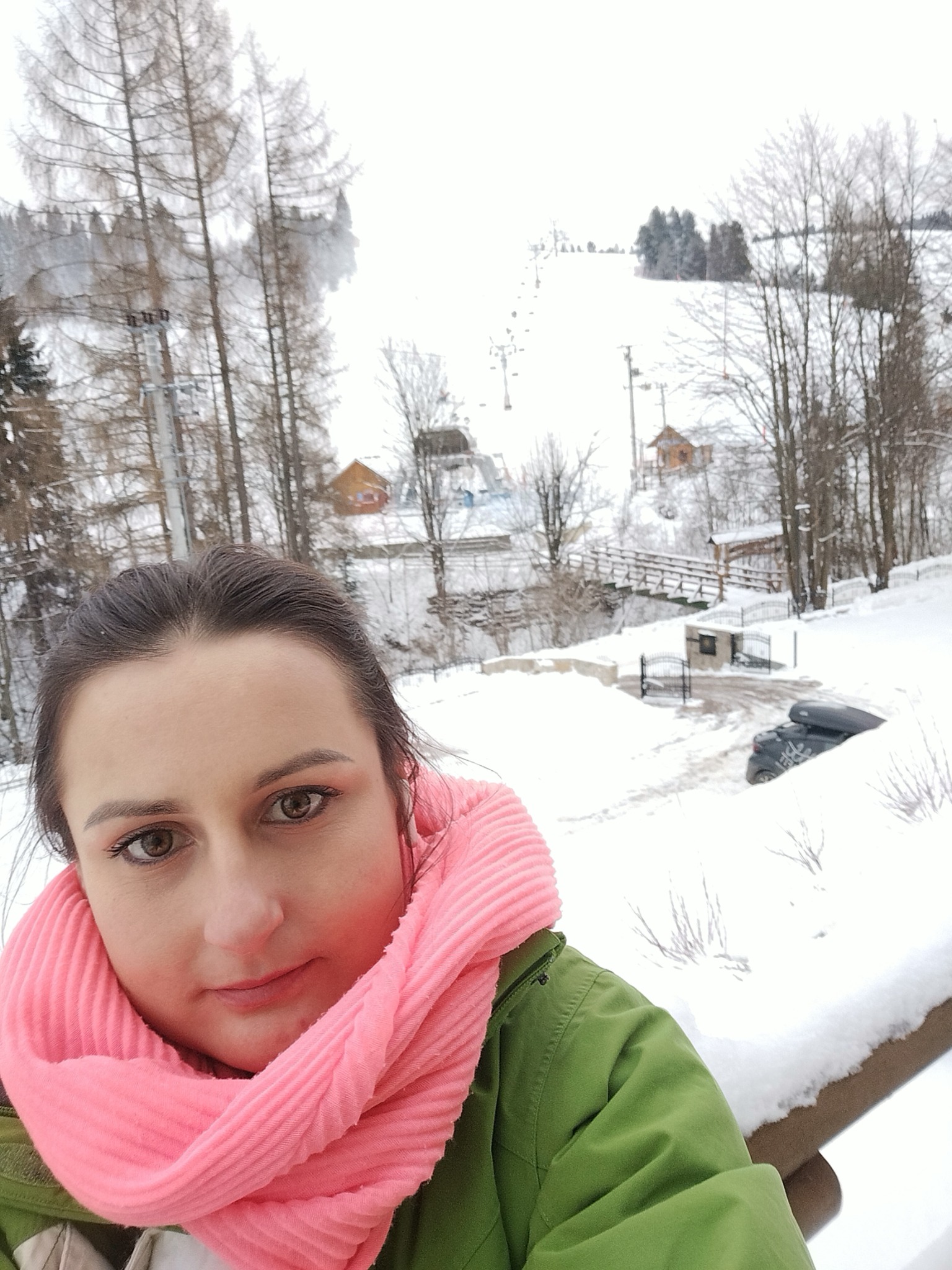 Kobieta w różowym szaliku i zielonej kurtce robiąca selfie na tle zimowego krajobrazu z zalesionymi wzgórzami i wyciągiem narciarskim w oddali.