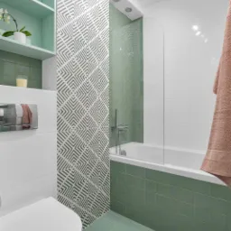 Wykończona łazienka w Warszawie z turkusowymi płytkami podłogowymi i częściowo na ścianach, białą armaturą i ścianą z geometrycznym wzorem. Nad toaletą zamontowane półki w kolorze płytek.