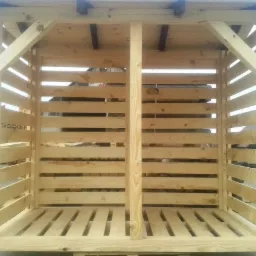 Surowa, drewniana konstrukcja z listew, prawdopodobnie składzik na drewno, z widoczną strukturą drewna i elementami łączeń, ustawiona na zewnątrz w słoneczny dzień.