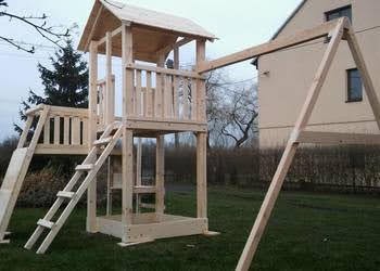 Surowa, drewniana konstrukcja placu zabaw z wieżą, zjeżdżalnią i huśtawką, w trakcie montażu na trawniku przed domem.