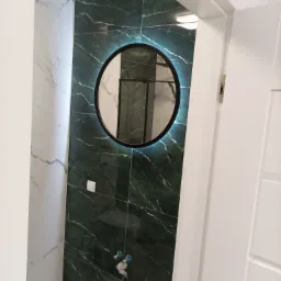 Wykończona łazienka z zielonymi płytkami imitującymi marmur, okrągłe lustro z podświetleniem LED oraz białe drzwi z minimalistycznym designem.