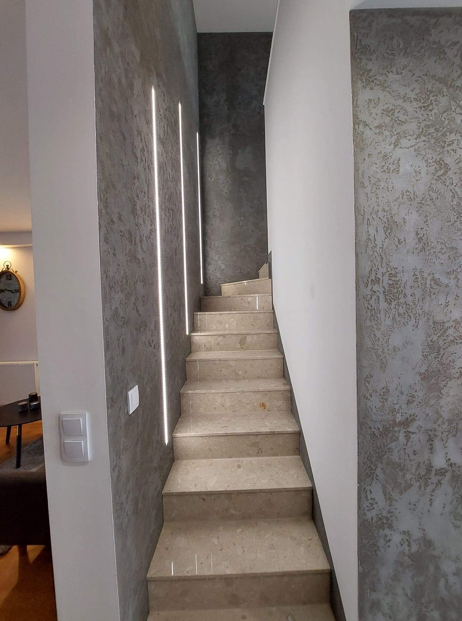 Klatka schodowa z marmurowymi stopniami, ściany wykończone w odcieniach szarości z dekoracyjną fakturą i wbudowanym oświetleniem LED.