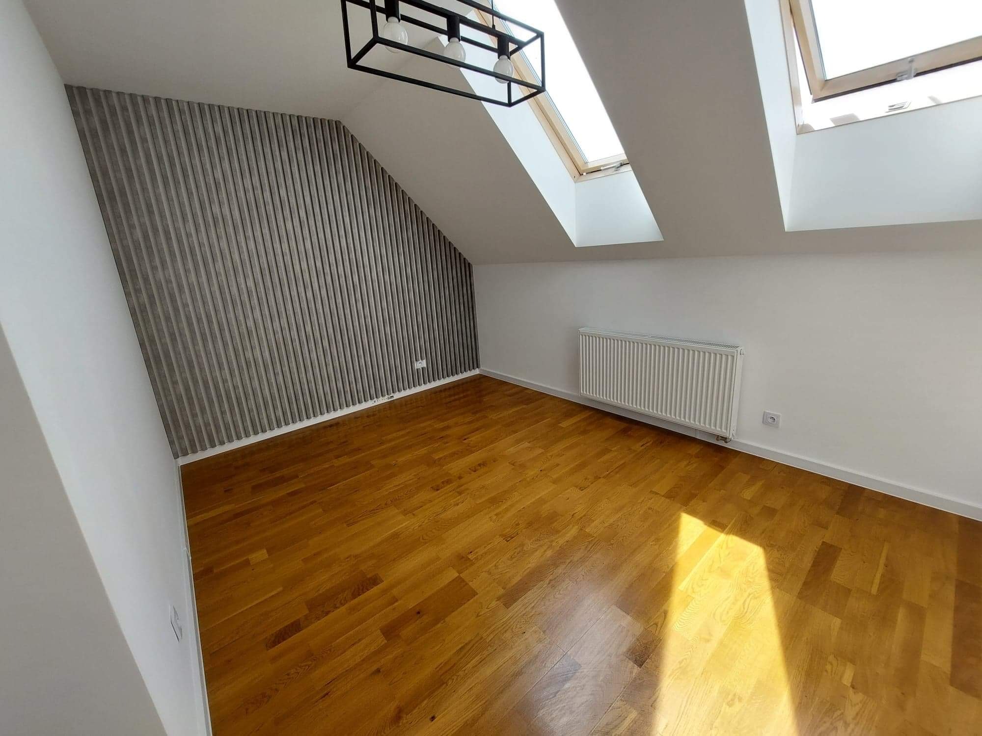 Poddasze po remoncie z drewnianą podłogą, grzejnikiem, oknami dachowymi i ścianą z dekoracyjnymi panelami pionowymi w odcieniach szarości.