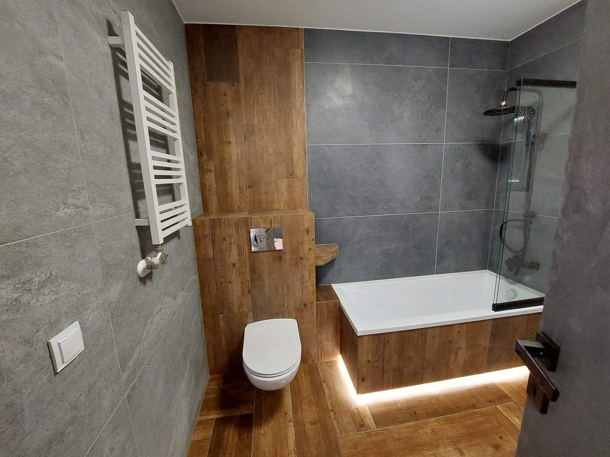 Nowoczesna łazienka z szarymi płytkami imitującymi kamień, drewnopodobnymi panelami i podświetlaną wanną, widoczny biały grzejnik drabinkowy i toaleta.