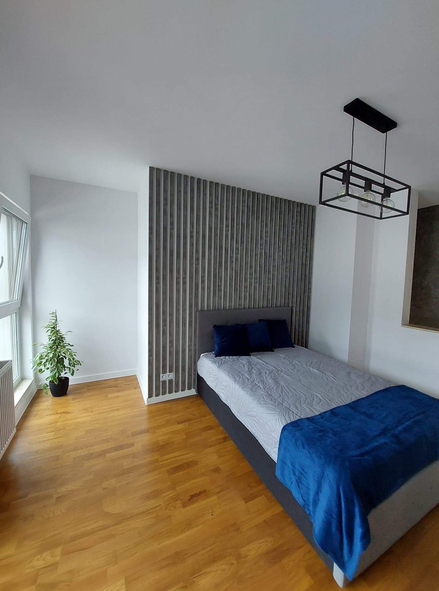 Nowoczesna sypialnia z drewnianą podłogą, szarą ścianą z pionowych paneli, łóżkiem z szarą ramą i niebieskimi poduszkami oraz wiszącą lampą z czarną, geometryczną oprawą.