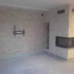 Salon w trakcie remontu z wykończoną ścianą z imitacji kamienia, zamontowanym uchwytem TV, przygotowanym miejscem na kominek i ułożonymi szczapami drewna.