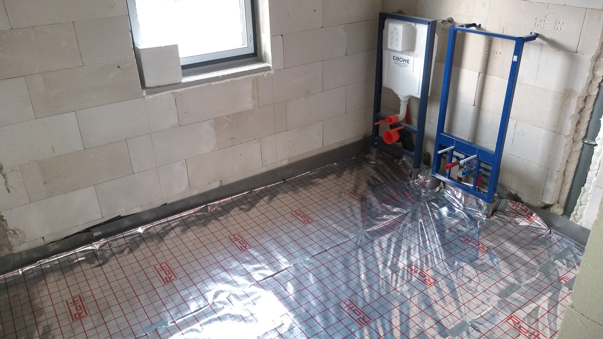 Łazienka w trakcie remontu: widoczne instalacje podtynkowe Grohe i mata grzewcza Roth na podłodze z bloczków betonowych.