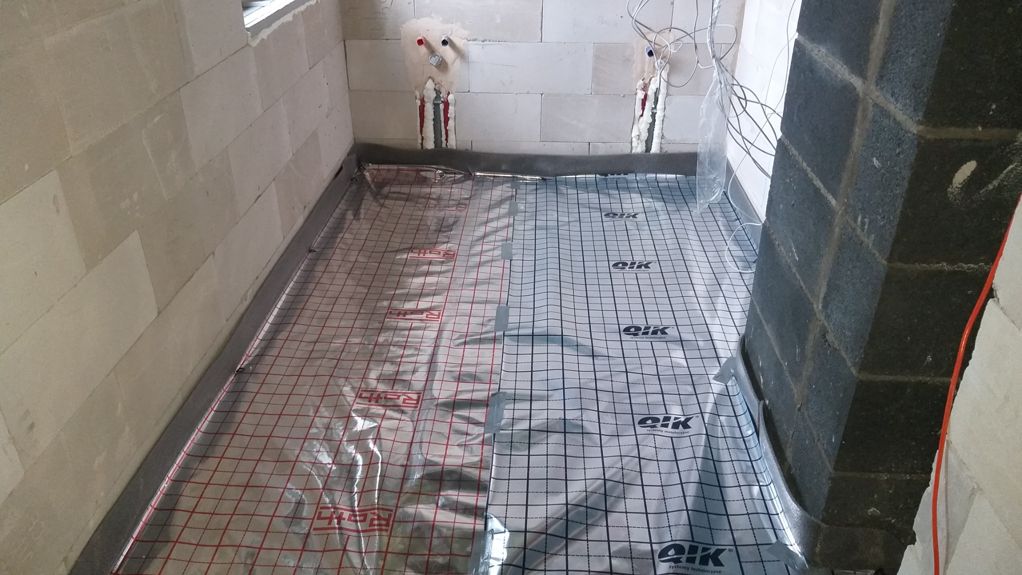Przygotowanie do montażu ogrzewania podłogowego: folia refleksyjna z siatką rastrową Roth i QIK na podłożu w surowej łazience, widoczne wyprowadzenia instalacji hydraulicznej.