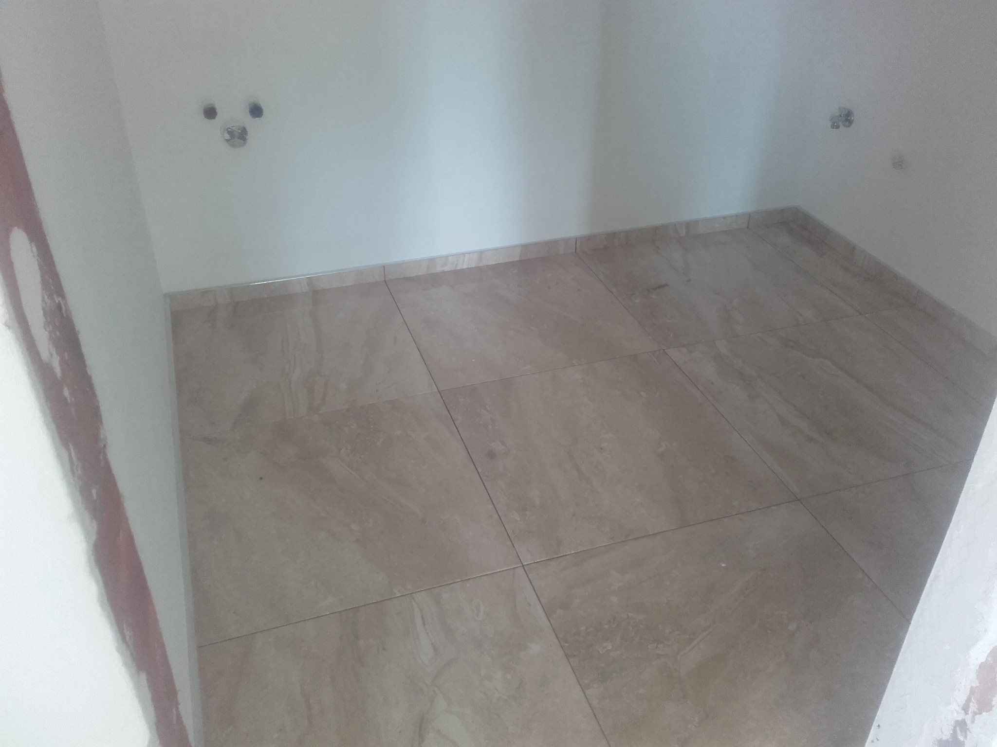 Wyłożona beżowymi płytkami podłoga w łazience w trakcie remontu, widoczne wyprowadzenia instalacji na ścianie, ślady po pracach wykończeniowych na ścianach.