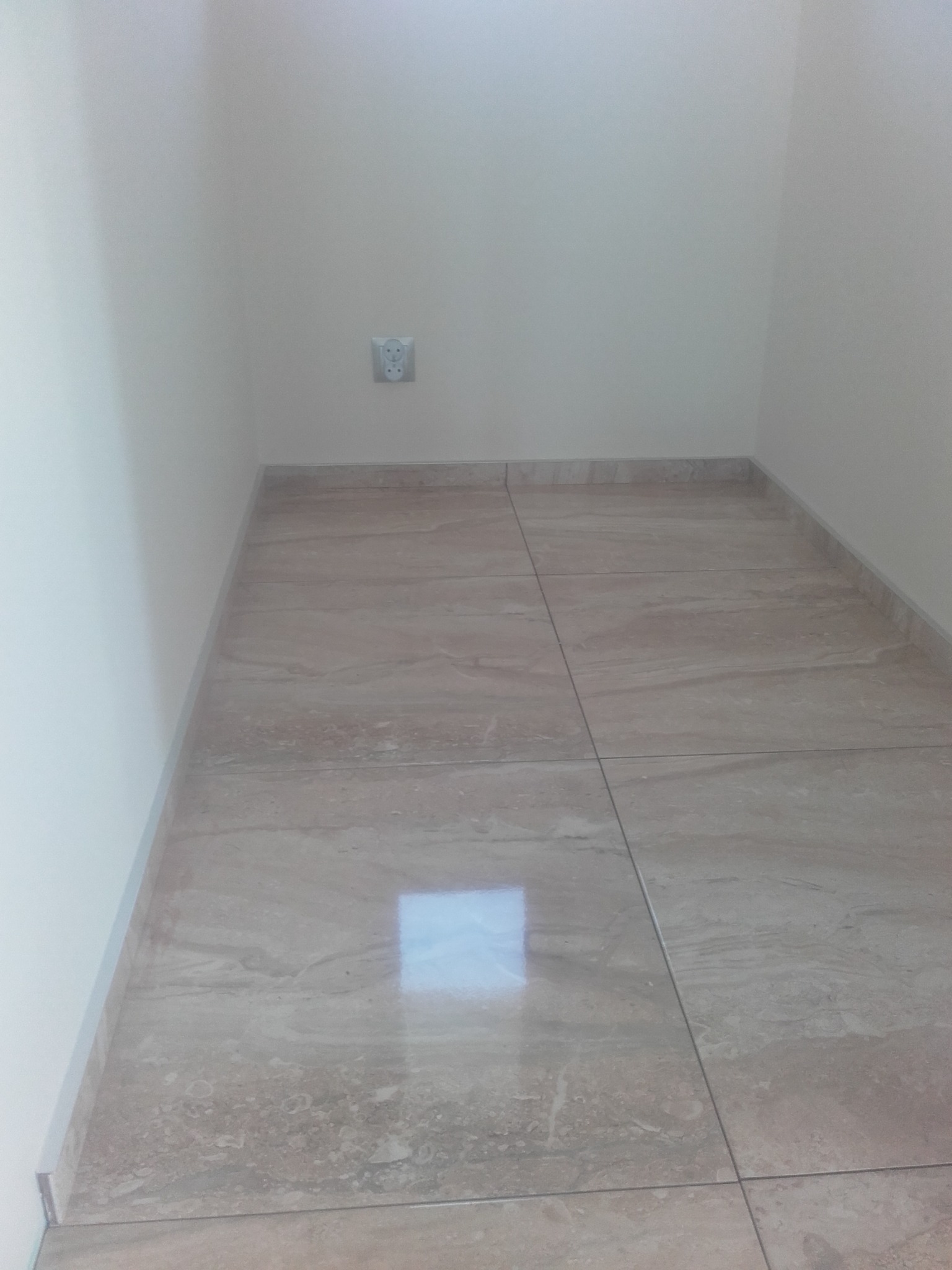 Wykończona podłoga w małej łazience z beżowymi płytkami imitującymi marmur, widoczne gniazdko elektryczne na białej ścianie.